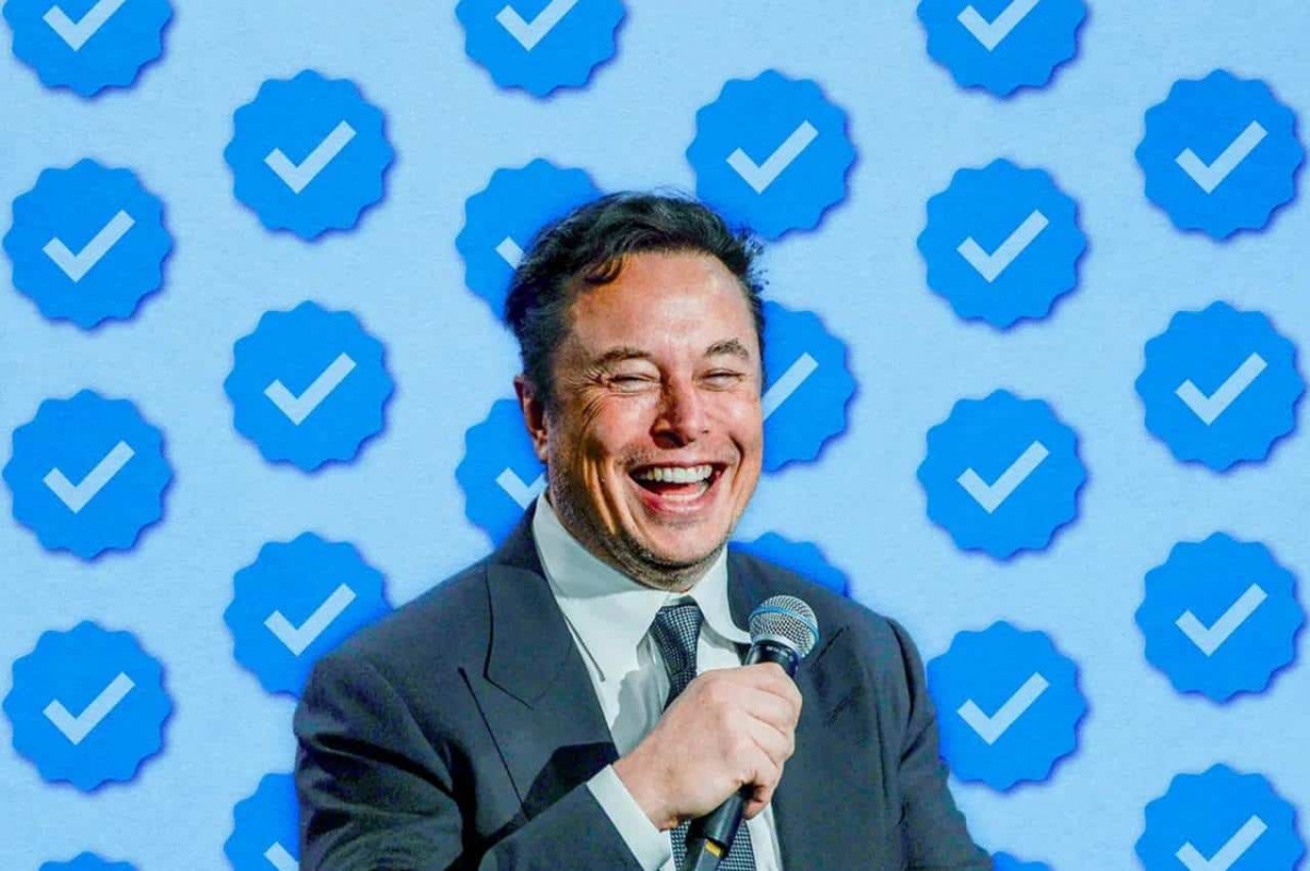 Elon Musk sorride, dietro di lui uno sfondo tappezzato di spunte blu